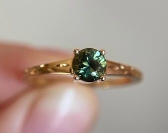 May include: A gold ring with a round, deep green gemstone. The band has a delicate, textured design. The gemstone is held in place by four prongs. The ring is a piece of jewelry.