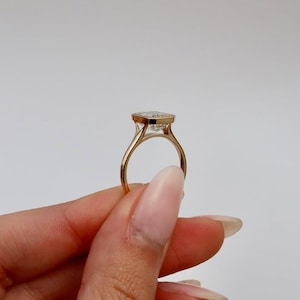 K&ouml;nnte beinhalten: Ein goldener Ring mit einem gro&szlig;en, quadratisch geschliffenen, klaren Edelstein. Der Ring hat ein zartes Band und eine L&uuml;nettenfassung. Der Ring wird vor einem schlichten wei&szlig;en Hintergrund gehalten.