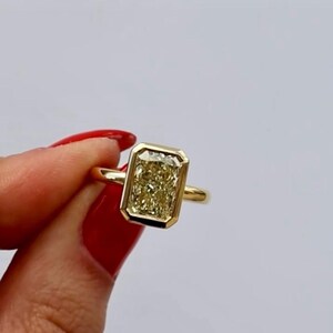 May include: A gold ring featuring a rectangular, yellow gemstone. The gemstone is set in a bezel setting, and the ring has a simple, polished band. The ring is held by a person with red nail polish.