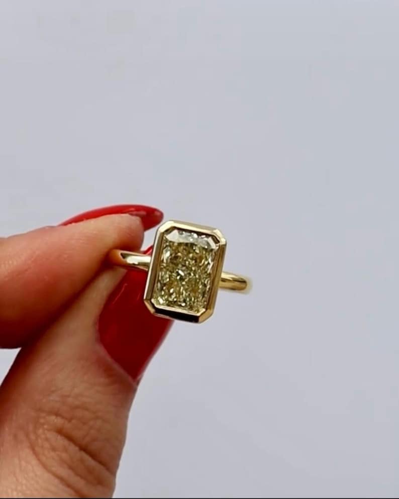 May include: A gold ring featuring a rectangular, yellow gemstone. The gemstone is set in a bezel setting, and the ring has a simple, polished band. The ring is held by a person with red nail polish.
