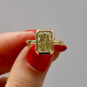 May include: A gold ring featuring a rectangular, yellow gemstone. The gemstone is set within a gold bezel, and the band is a simple, polished gold. The ring is held up against a neutral background.