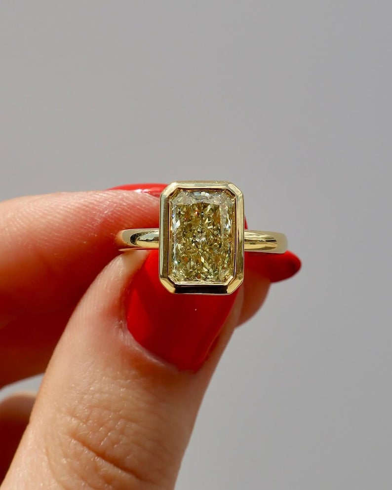 May include: A gold ring featuring a rectangular, yellow gemstone. The gemstone is set within a gold bezel, and the band is a simple, polished gold. The ring is held up against a neutral background.