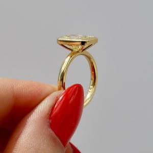 May include: A gold ring with a rectangular, light-colored gemstone. The ring's band is thin and polished, and the gemstone is set in a bezel setting. The ring is held by a person with red nail polish.