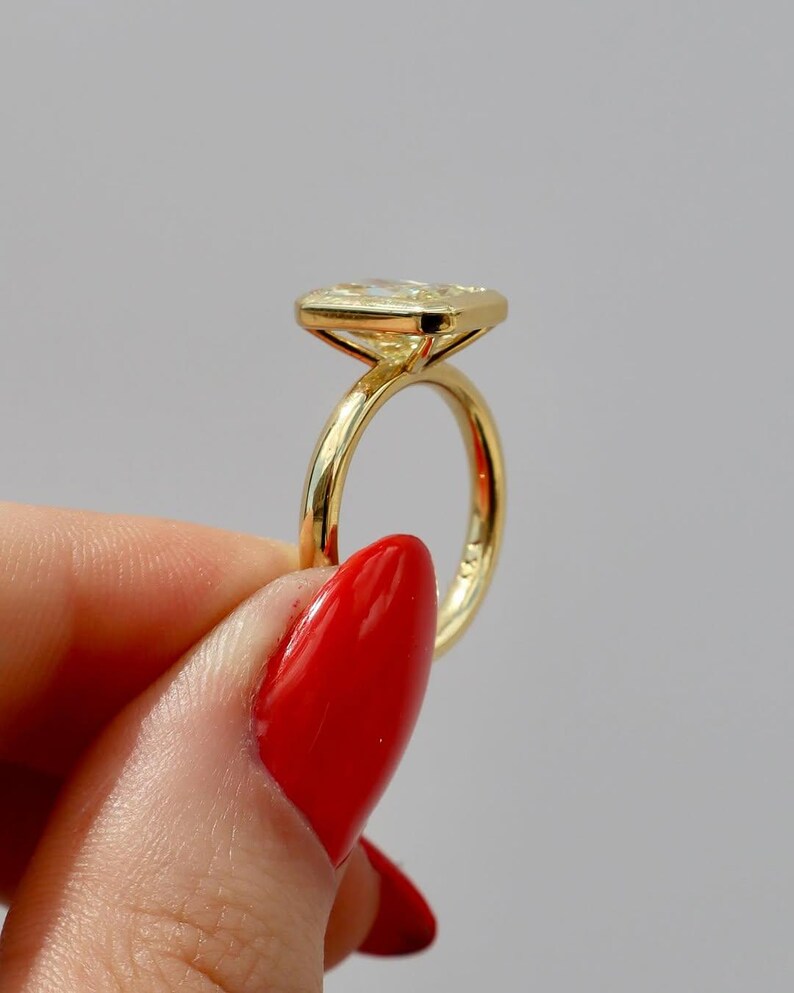 May include: A gold ring with a rectangular, light-colored gemstone. The ring's band is thin and polished, and the gemstone is set in a bezel setting. The ring is held by a person with red nail polish.