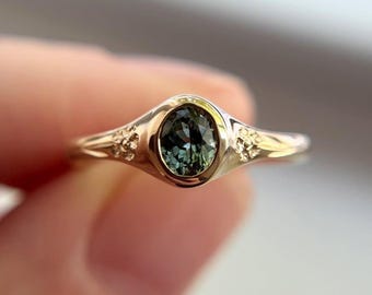 May include: A gold ring with an oval, teal-colored gemstone. The ring has a delicate design with small floral accents on either side of the gemstone. The band is thin and polished, creating an elegant and refined look.