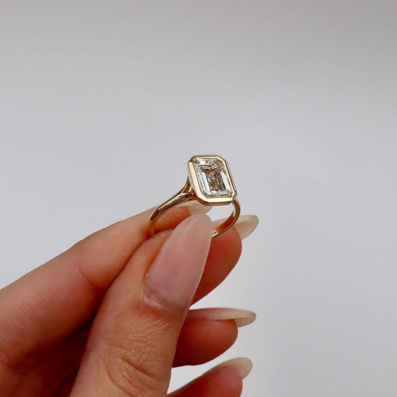 K&ouml;nnte beinhalten: Ein goldener Ring mit einem Diamanten im Smaragdschliff. Der Diamant ist in einer L&uuml;nettenfassung gefasst, und das Band ist aus poliertem Gold. Der Ring wird vor einem wei&szlig;en Hintergrund gezeigt.
