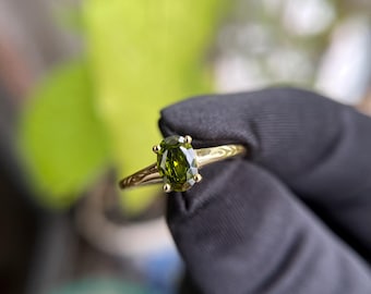 May include: A gold ring featuring an oval-cut, olive-green gemstone. The ring's band has a delicate, engraved design. The ring is held against a blurred green background, with a black glove in the foreground.