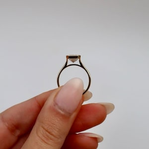 K&ouml;nnte beinhalten: Ein goldener Ring mit einem quadratisch geschliffenen, klaren Edelstein. Der Ring wird vor einem wei&szlig;en Hintergrund gehalten, wodurch das zarte Band und die Fassung des Steins hervorgehoben werden. Das Design des Rings ist schlicht und elegant.