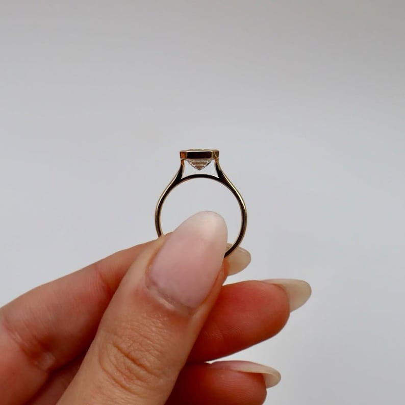 K&ouml;nnte beinhalten: Ein goldener Ring mit einem quadratisch geschliffenen, klaren Edelstein. Der Ring wird vor einem wei&szlig;en Hintergrund gehalten, wodurch das zarte Band und die Fassung des Steins hervorgehoben werden. Das Design des Rings ist schlicht und elegant.
