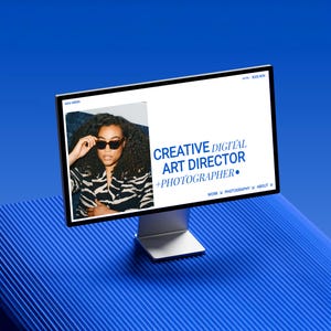 May include: A computer monitor displays a website with the text "CREATIVE DIGITAL ART DIRECTOR + PHOTOGRAPHER." The website features a photo of a person wearing sunglasses and a black and white patterned top. The monitor is on a silver stand against a blue background.
