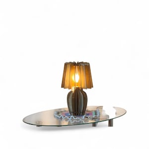 Könnte beinhalten: Eine Tischlampe aus Holz mit braunem Finish und einem einzigartigen Schirmdesign. Die Lampe steht auf einem Glastisch mit Metallbeinen, darunter eine bunte Tischdecke. Die Lampe ist beleuchtet und strahlt ein warmes Licht aus.