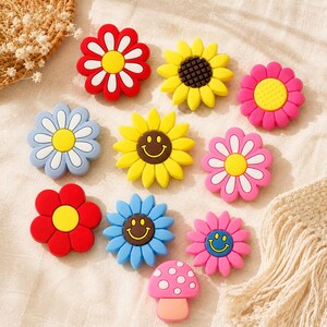 May include: A collection of ten colorful silicone charms featuring various flower shapes and one mushroom. The designs include red, yellow, pink, and blue petals with yellow or brown centers, and three of the flowers feature a yellow smiley face.
