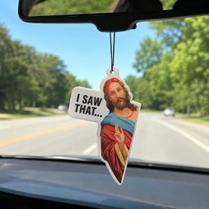 May include: A car air freshener hanging from a rearview mirror, featuring a printed illustration of a bearded figure with long brown hair wearing a red and blue robe, with a speech bubble containing the black text I SAW THAT...