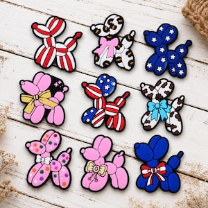 May include: A collection of nine balloon animal dog charms in various patterns and colours. Designs include red and white stripes, leopard print with a pink bow, blue with white stars, pink with a yellow bow, patriotic stripes and stars, cow print with a blue bow, pink with colourful stars, pink with a beige bow, and blue with a red, white, and blue bow.