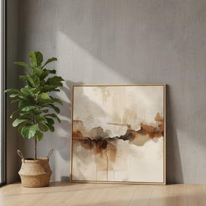 May include: A square framed abstract painting featuring layered washes of beige, cream, and deep brown tones. Beside the artwork stands a tall green fiddle leaf fig plant housed in a woven wicker basket with two small handles on the sides.