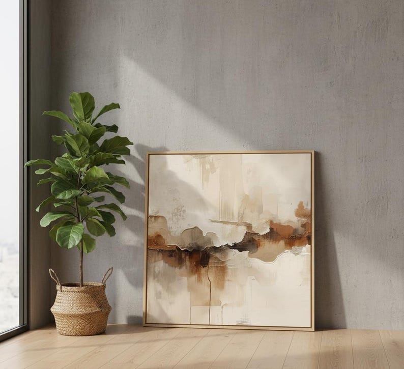 May include: A square framed abstract painting featuring layered washes of beige, cream, and deep brown tones. Beside the artwork stands a tall green fiddle leaf fig plant housed in a woven wicker basket with two small handles on the sides.