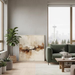 May include: A square framed wall art piece featuring an abstract landscape painting with layered washes of cream, beige, and deep brown tones. The artwork is displayed in a thin light-colored frame, showcasing fluid, organic shapes and textures.