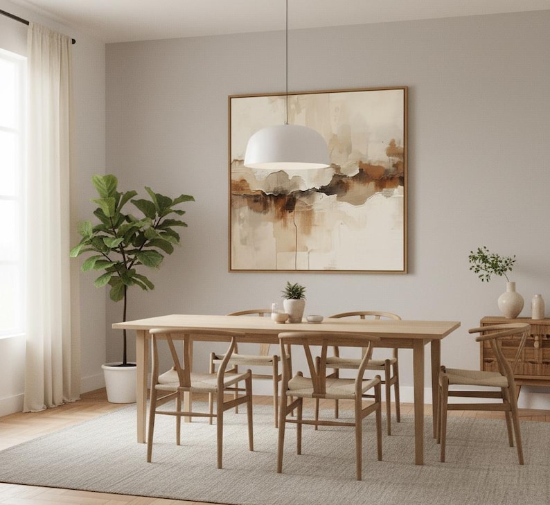 May include: A light wood dining table with four matching chairs featuring woven seats, positioned under a white dome pendant light. A large square abstract painting with beige, brown, and white tones hangs on the wall above the table.