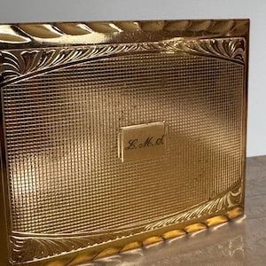 May include: A rectangular gold-tone metal case featuring a fine grid-patterned texture on the front surface. The center includes a smooth, raised rectangular plaque engraved with the cursive initials L.M.S. The edges have a decorative border.