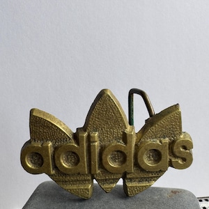 May include: A brass-toned metal belt buckle featuring the Adidas trefoil logo and the word "adidas" in raised, textured lettering. The buckle includes a metal prong on the back for attachment to a belt strap, showcasing a weathered, metallic finish.