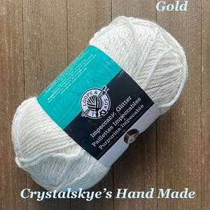 May include: A skein of off-white yarn with glitter, labeled "Impeccable Glitter." The label is black and teal, with the brand name "Loops & Threads." The text "Crystalskye's Hand Made" is at the bottom. The word "Gold" is in the top right corner.