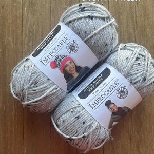 May include: Four skeins of gray tweed yarn with black flecks, labeled "Impeccable" with a woman wearing a hat. The yarn is on a wooden surface. The text "Brown Tweed" is in the upper right corner.