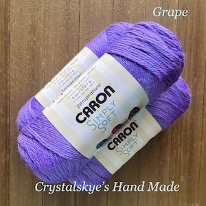 May include: Three skeins of Caron Simply Soft yarn in the color "Grape" are displayed. Each skein is labeled with the brand name, weight of 6 oz (170.1 g), and fiber content. The text "Crystalskye's Hand Made" is at the bottom.