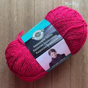 May include: A vibrant ruby-colored yarn ball with glitter accents. The label reads "Loops & Threads Impeccable Glitter". The yarn is 217 yards / 198 m. Ideal for knitting and crochet projects.