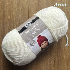 May include: A skein of off-white yarn labeled "Impeccable" by Loops & Threads. The label features a photo of a child wearing a red knit hat. The word "Aran" is printed in the upper right corner.
