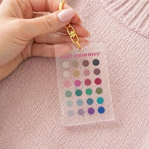 May include: A clear acrylic keychain with a gold clasp and the words "soft summer" printed in pink. The keychain features a grid of small, colorful dots in various shades, held against a pink sweater.
