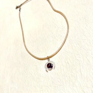 Puede incluir: Un delicado collar con un cordón color crema y un cierre plateado. El colgante es un dije transparente en forma de media luna con una gema de color púrpura oscuro en el centro. El collar se muestra sobre una superficie texturizada blanquecina.