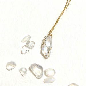 Puede incluir: Un collar con colgante de cristal de cuarzo transparente con una cadena y un alambre dorado. Varias piedras de cuarzo en bruto están dispersas alrededor del collar. El collar es una joya delicada.