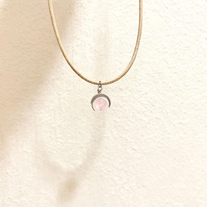 Puede incluir: Un delicado collar con un pequeño colgante de piedra rosa en un engaste plateado. El colgante está suspendido de un fino cordón beige. El collar se muestra sobre un fondo liso blanquecino.