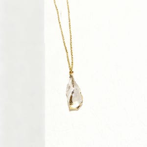 Op de afbeelding: Een goudkleurige ketting met een heldere, onregelmatig gevormde hanger. De hanger heeft goudkleurige accenten langs de randen. De ketting wordt gepresenteerd tegen een witte achtergrond.