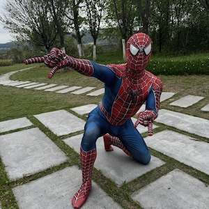 May include: A person in a Spider-Man costume, kneeling on a stone surface. The costume is red and blue with a web pattern. The person is outdoors with trees in the background.