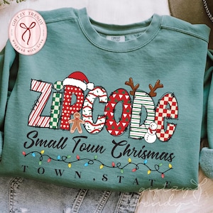 May include: A teal sweatshirt with the text "ZIPCODE Small Town Christmas" in a festive design. The word "ZIPCODE" features Christmas-themed patterns and holiday accents, including a Santa hat, reindeer antlers, and a snowman.