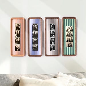 May include: Four rectangular wooden frames mounted on a wall, each containing a vertical strip of black and white photo booth pictures. The frames feature striped fabric backings in shades of orange, blue, grey, and green, respectively.