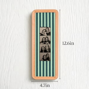 Puede incluir: Un marco de madera rectangular que contiene una tira vertical de cuatro fotos en blanco y negro de dos personas. El fondo detr&aacute;s de las fotos tiene rayas verticales de color verde oscuro y verde claro. Las dimensiones son 32cm por 12cm.