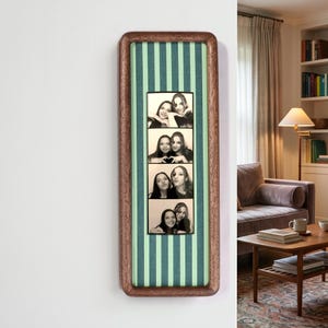 Puede incluir: Un marco de madera rectangular vertical con esquinas redondeadas, que presenta un fondo de tela a rayas de color verde claro y verde oscuro. Centrada en el interior hay una tira de fotos en blanco y negro que muestra cuatro poses de dos personas.