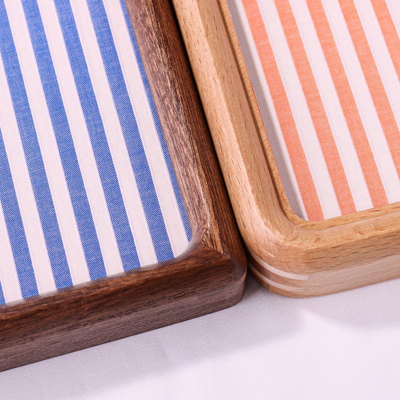 Puede incluir: Dos bandejas rectangulares de madera con esquinas redondeadas. Una bandeja presenta un marco de madera marr&oacute;n oscuro con una incrustaci&oacute;n de tela a rayas verticales azules y blancas. La segunda bandeja tiene un marco de madera clara con rayas naranjas.