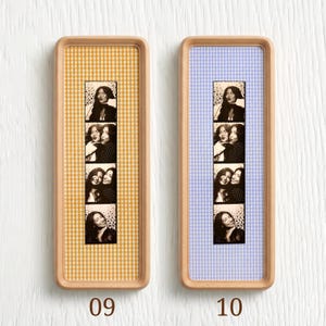 Puede incluir: Dos marcos rectangulares de madera que contienen tiras de fotos verticales. El marco izquierdo presenta un fondo de cuadros vichy amarillos y blancos, mientras que el derecho tiene un fondo de cuadros vichy azul claro y blancos. Cada tira muestra cuatro fotos en blanco y negro.