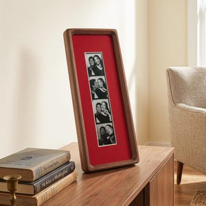 Può includere: Una cornice fotografica rettangolare in legno con interno rosso contiene una striscia di foto in bianco e nero. La cornice è in legno scuro. Le foto mostrano coppie. La cornice è alta circa 30 cm e si trova su una superficie di legno.