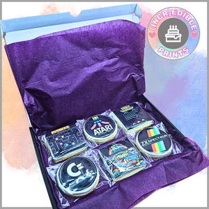 May include: A presentation box containing iced biscuits decorated with retro video game imagery. The biscuits are individually wrapped and presented on purple tissue paper. The box is open, revealing the treats inside. The box has the text "(INCREDIBLE) PRINTS" on the top right.