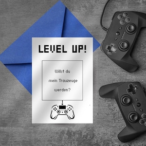 Puede incluir: Una tarjeta blanca con el texto "LEVEL UP!" y "Willst du mein Trauzeuge werden?" está junto a un sobre azul y dos mandos de juego negros. La tarjeta presenta un gráfico de mando.