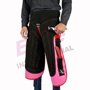 May include: Black and pink leather farrier chaps with red stitching and trim. The chaps have pockets and tool holders. A person is holding a farrier's hammer.