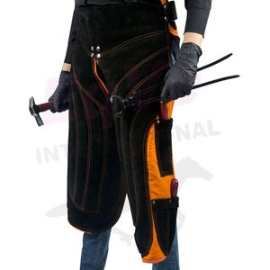 May include: Black and orange leather farrier apron with orange stitching. The apron has pockets and is worn by a person wearing black gloves. A hammer and farrier tools are visible.