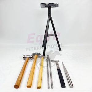 Pode incluir: Uma coleção de ferramentas de ferrador, incluindo martelos com cabos de madeira, uma faca para cascos e um suporte de metal. As ferramentas estão dispostas sobre uma superfície branca. O suporte é preto.