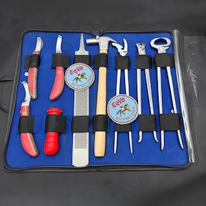 Puede incluir: Un estuche azul con cremallera que contiene un juego de herramientas de herrador. Las herramientas incluyen cuchillos para cascos, una escofina, un martillo, tenazas y otros instrumentos. Cada herramienta está asegurada con una correa negra. El estuche tiene dos parches circulares con el texto "Eqve International".