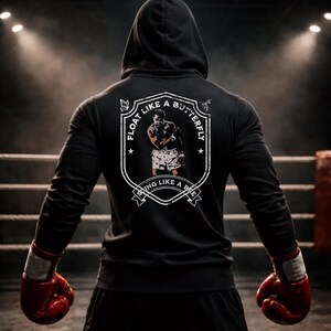 Könnte beinhalten: Schwarzer Kapuzenpullover mit dem Text "FLOAT LIKE A BUTTERFLY, STING LIKE A BEE" und einem Boxer-Bild. Die Person trägt rote Boxhandschuhe und steht in einem Boxring.