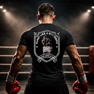 Könnte beinhalten: Schwarzes T-Shirt mit einem Aufdruck auf dem Rücken, der einen Boxer und den Text "Float Like a Butterfly, Sting Like a Bee" zeigt. Der Boxer trägt Shorts und Handschuhe. Das T-Shirt wird von einer Person in einem Boxring getragen.